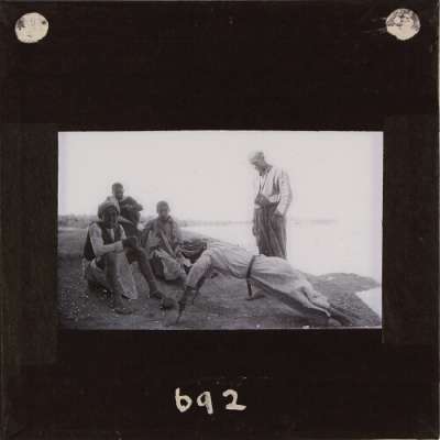 Lantern Slide: Men and boys by river shore, one doing press-up exercise